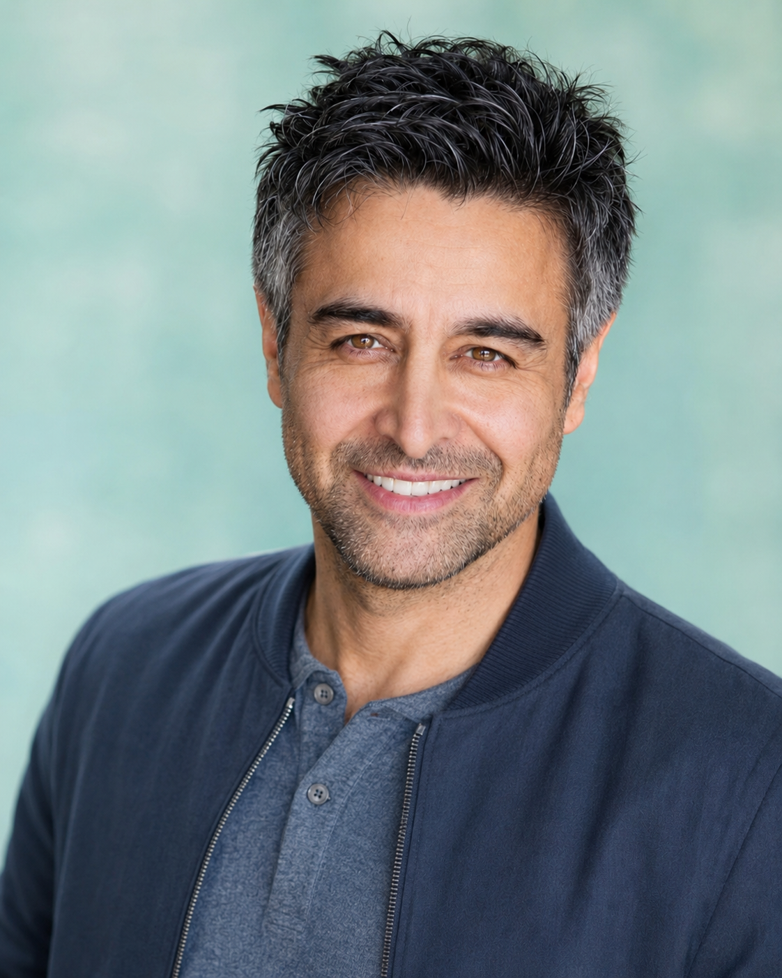 Nicholas Guilak — navy bomber jacket on a turquoise backdrop, warm smile